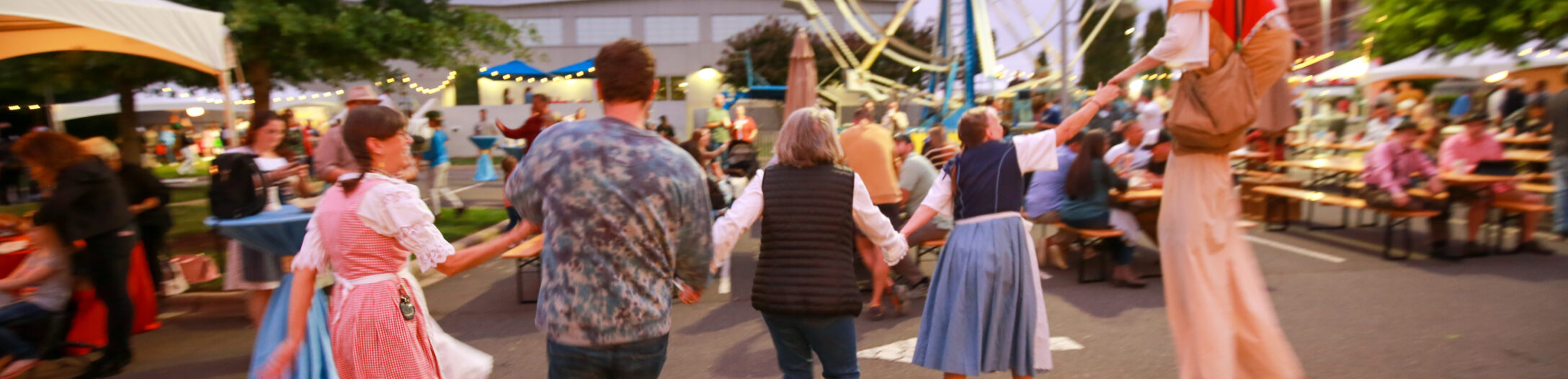 People in dirndl and lederhosen dance with a stilt walker at a carnival at dusk. There is a ferris wheel in the background.